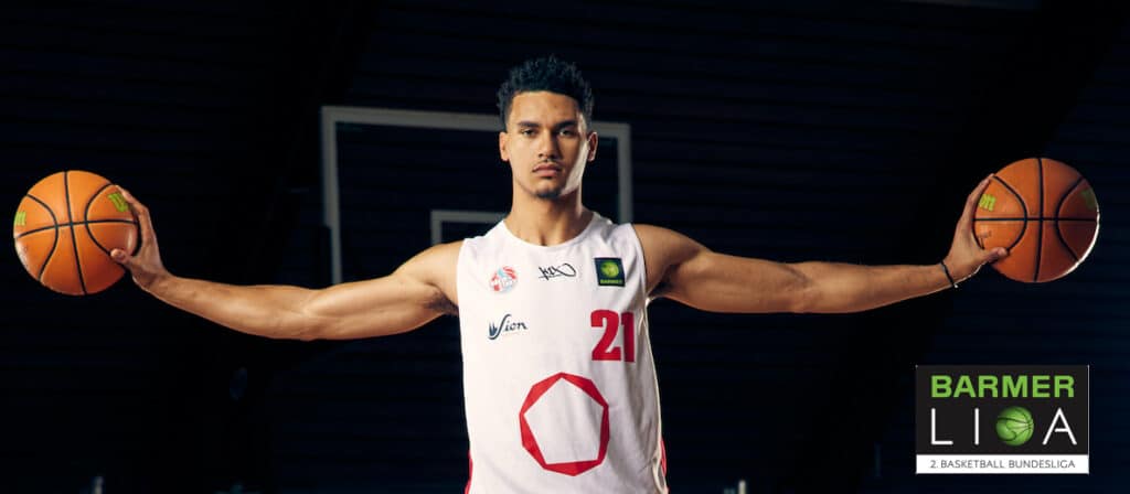 Basketballspieler in weißem Trikot Nummer 21 hält zwei Basketbälle mit ausgestreckten Armen.