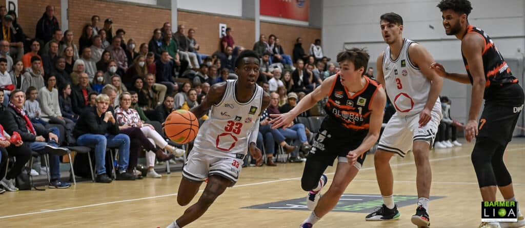 Basketballspiel der RheinStars Köln: Spieler im weißen Trikot dribbelt gegen Gegner im schwarzen Trikot mit Zuschauern im Hintergrund.