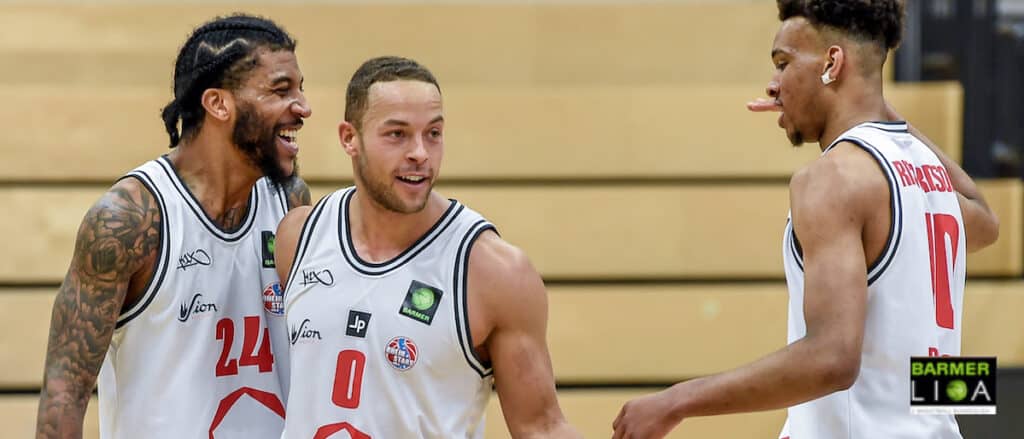 Drei Basketballspieler der RheinStars Köln lachen zusammen auf dem Spielfeld.