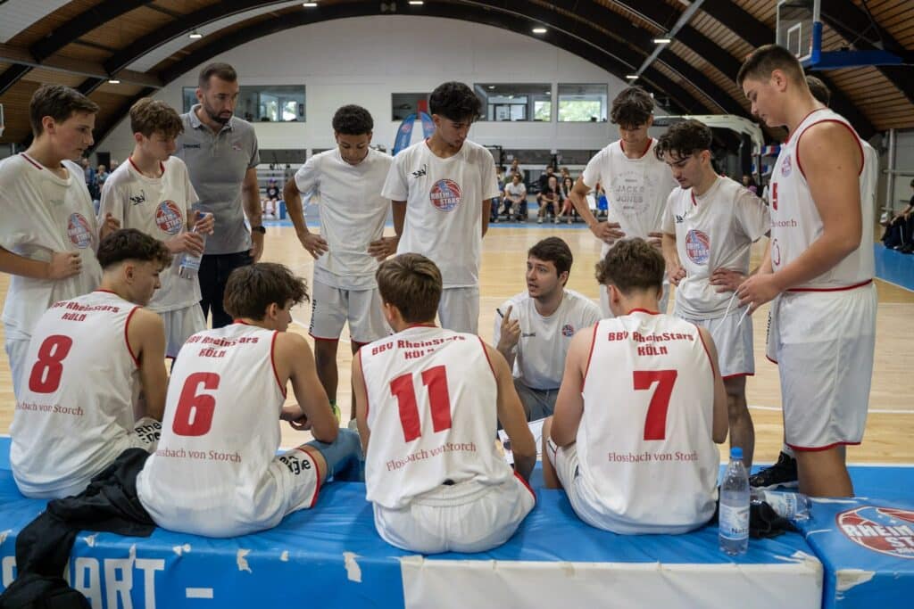 Basketballmannschaft RheinStars Köln in Trainingspause, Coach spricht zu sitzenden Spielern in Sporthalle.