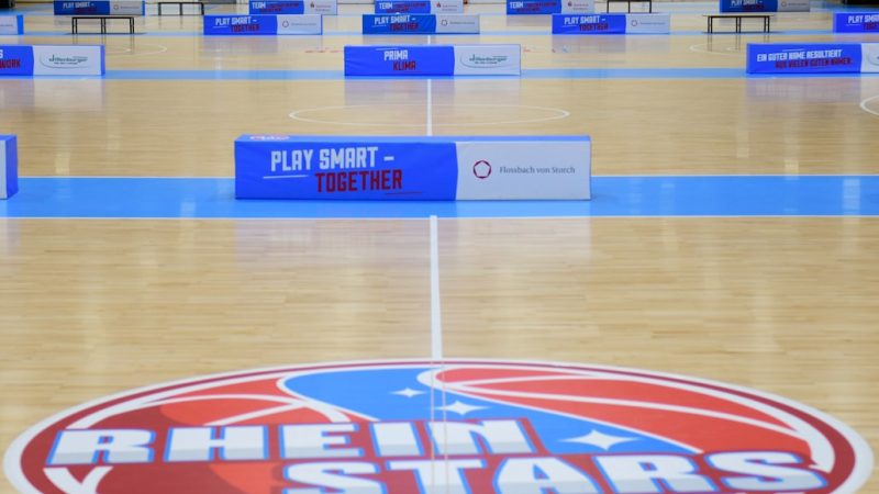 Basketballhalle der RheinStars Köln mit Teamlogo auf dem Parkettboden und blauen Werbebanden.