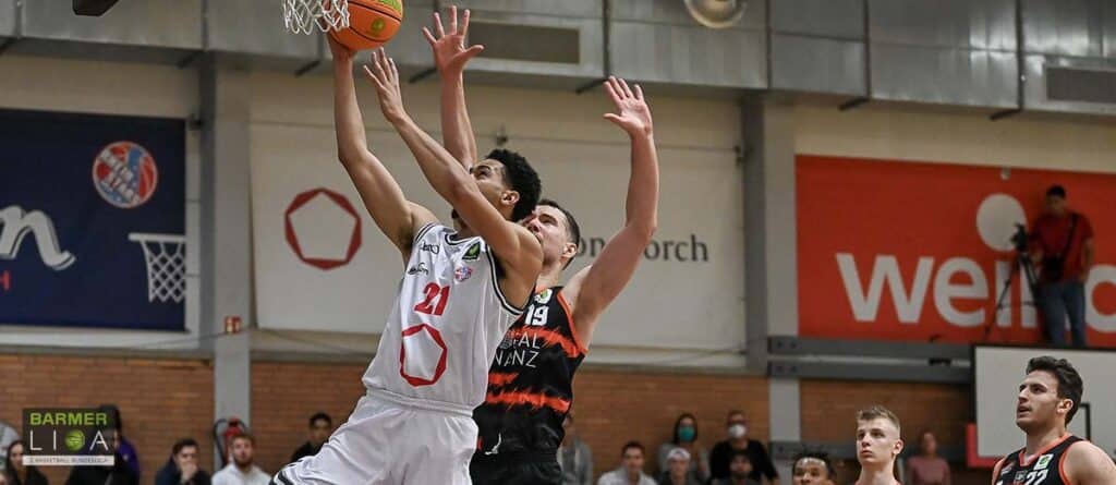 Basketballspiel der RheinStars Köln: Spieler in Weiß springt zum Wurf, Gegenspieler in Schwarz-Rot verteidigt.