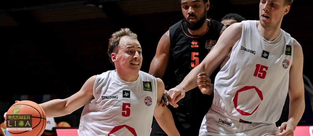 Basketballspieler der RheinStars Köln in weißen Trikots während eines Spiels gegen schwarze Gegner.