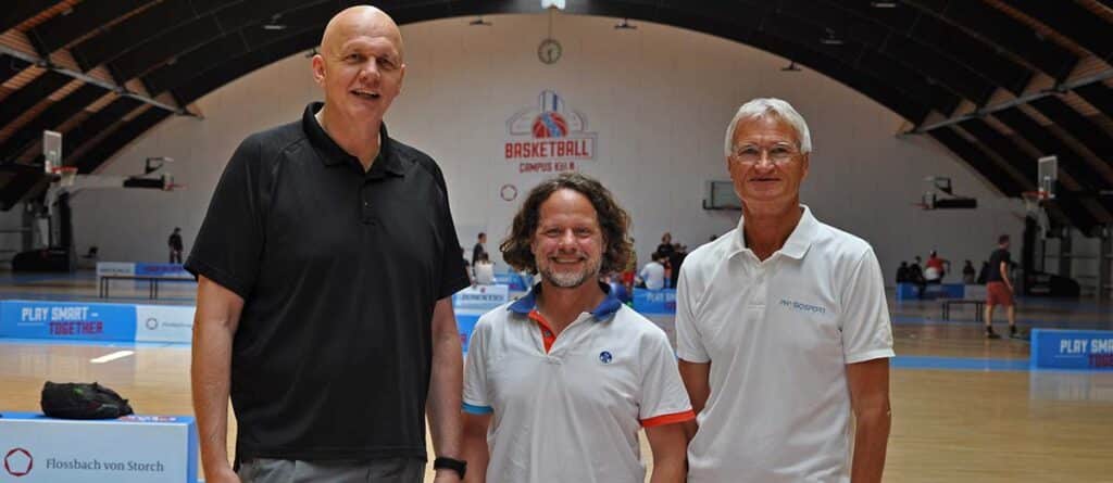 Drei Männer stehen in einer Basketballhalle der RheinStars Köln nebeneinander.