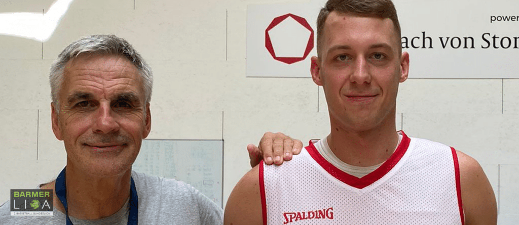 Zwei Männer posieren zusammen, einer trägt ein rotes und weißes Spalding-Basketballtrikot der RheinStars Köln.