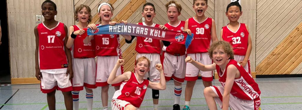 Jubelnd posiert die Jugendmannschaft der RheinStars Köln in roten Basketballtrikots mit einem Schal in einer Sporthalle.