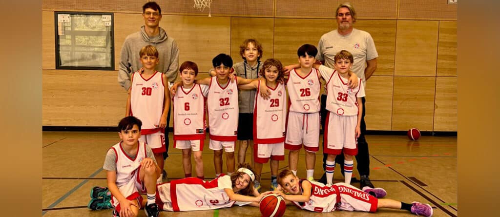 Basketballmannschaft RheinStars Köln posiert in der Sporthalle mit Trainern und Spielern in roten und weißen Trikots.