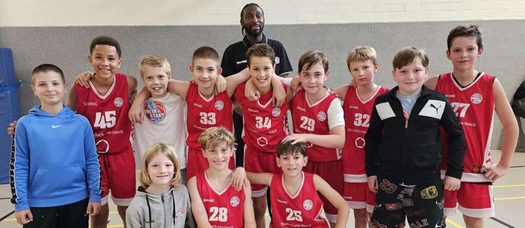 Jugendbasketballmannschaft der RheinStars Köln posiert zusammen mit Trainer in einer Sporthalle.