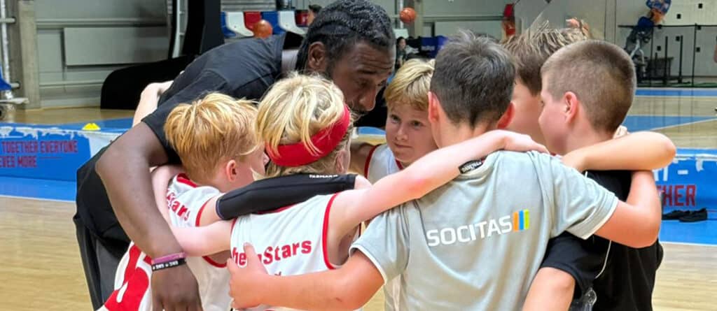 Ein Trainer der RheinStars Köln motiviert junge Spieler in einer Mannschaftsumarmung in der Sporthalle.