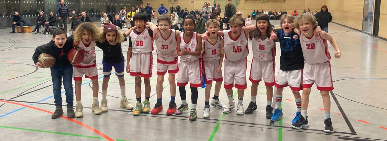 Jugendbasketballteam der RheinStars Köln posiert lachend in einer Sporthalle.