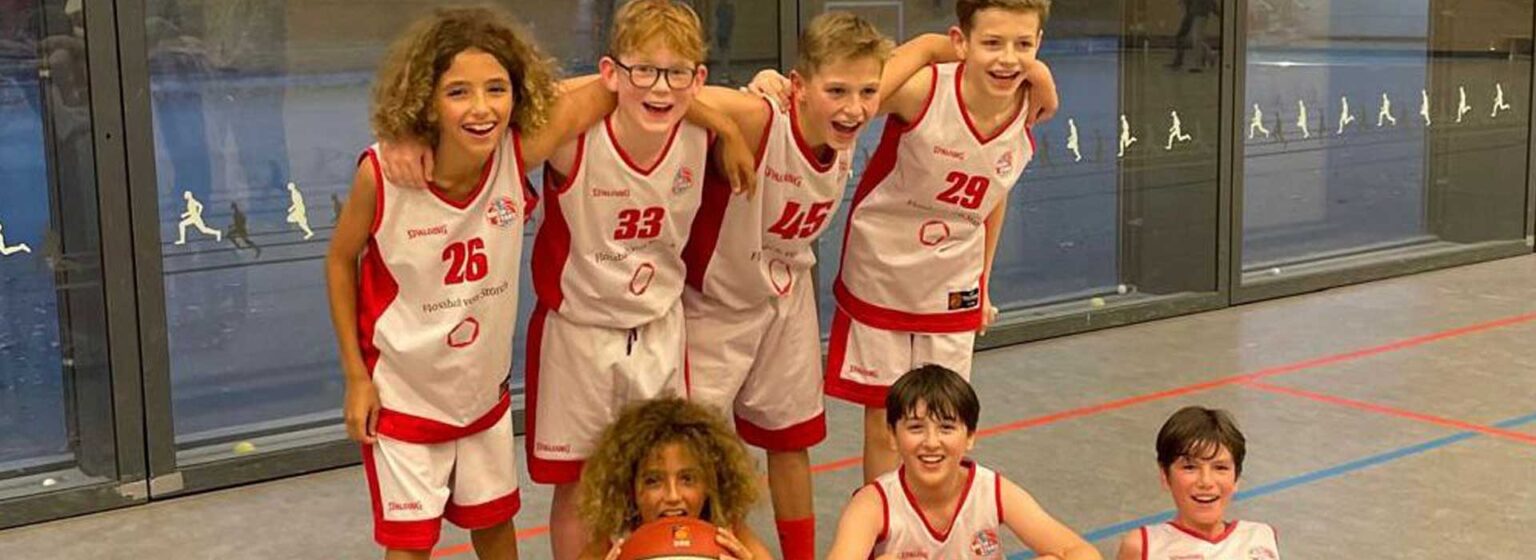 Junge Basketball-Mannschaft der RheinStars Köln posiert lachend in rot-weißen Trikots in einer Sporthalle.