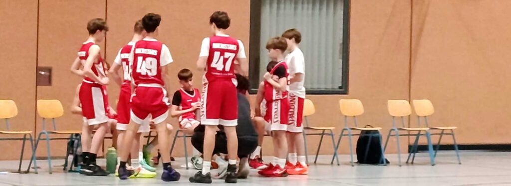 Jugendbasketballteam RheinStars Köln in roten Trikots während Auszeit in Sporthalle.