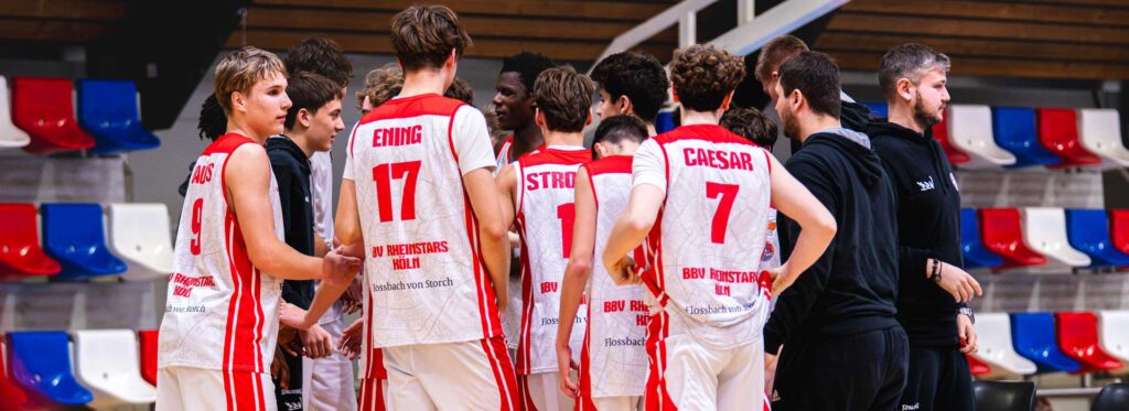 Basketballmannschaft RheinStars Köln im Teamkreis mit Trainerstab in der Halle.
