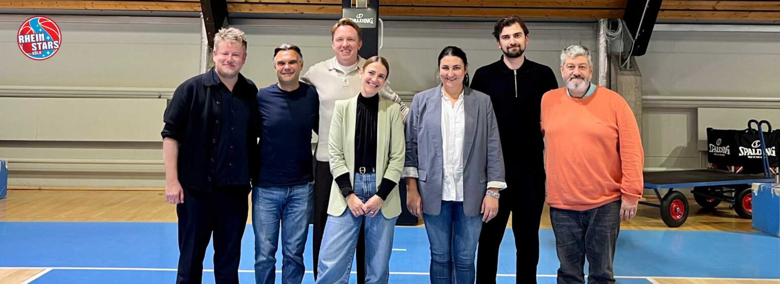 Sieben Personen stehen in einer Sporthalle der RheinStars Köln und posieren für ein Gruppenfoto.