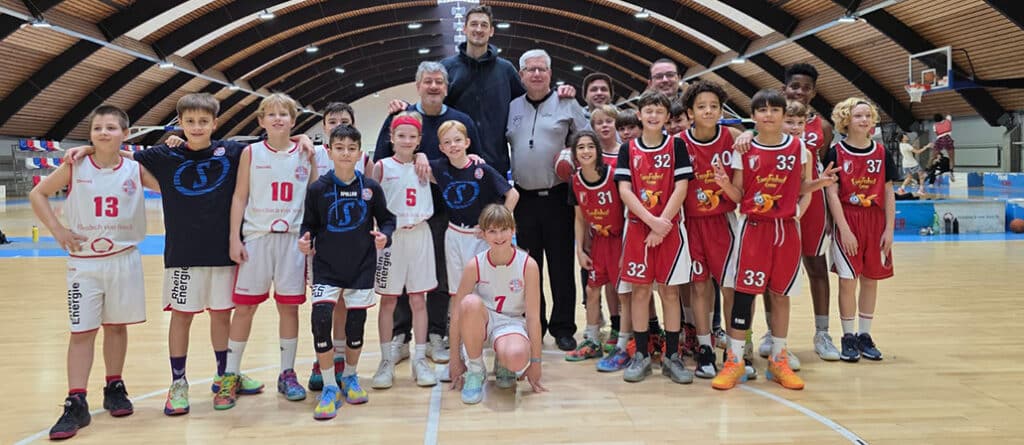 Zwei Kinder-Basketballmannschaften der RheinStars Köln posieren zusammen mit Trainern in einer Sporthalle.