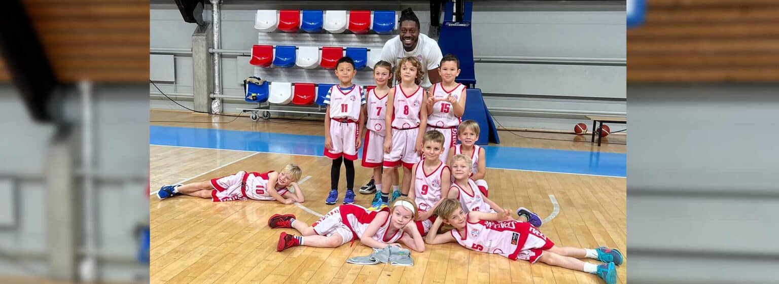 Kinder-Basketballteam der RheinStars Köln posiert spielerisch auf dem Hallenboden mit Trainer.