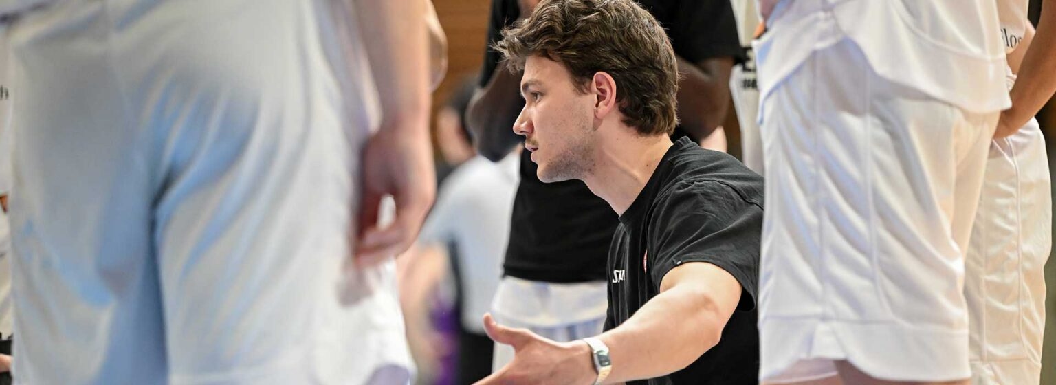 Mann in schwarzem Shirt gestikuliert während Trainingseinheit der RheinStars Köln, umgeben von Spielern in weißen Trikots.
