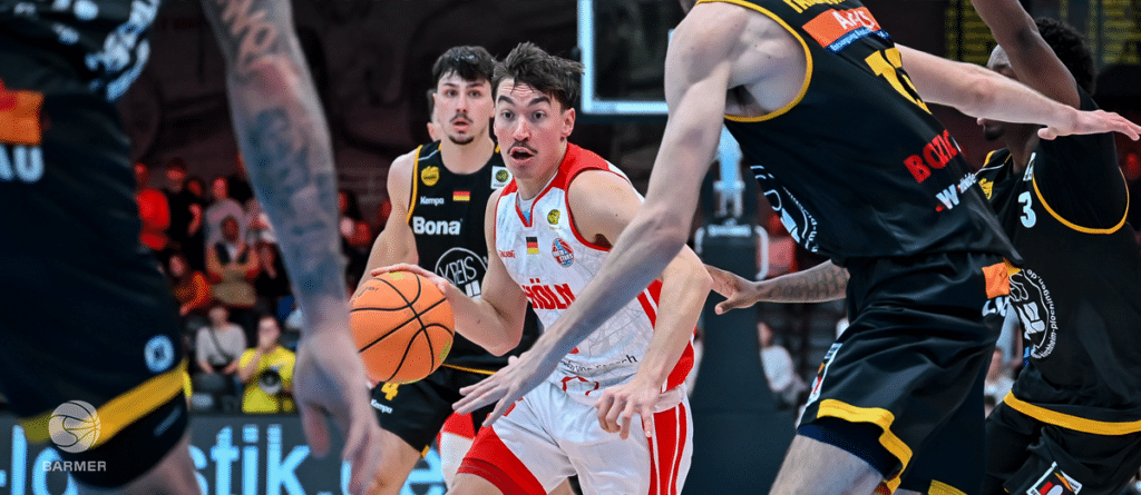 Basketballspiel: RheinStars-Spieler in Rot-Weiß dribbelt gegen schwarze Gegner.