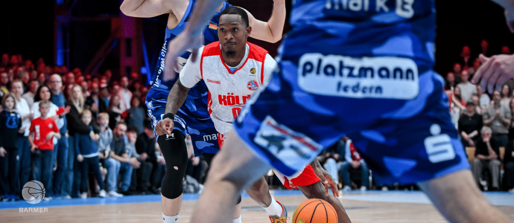 Basketballspieler der RheinStars Köln in rotem Trikot während eines Spiels gegen blaues Team.