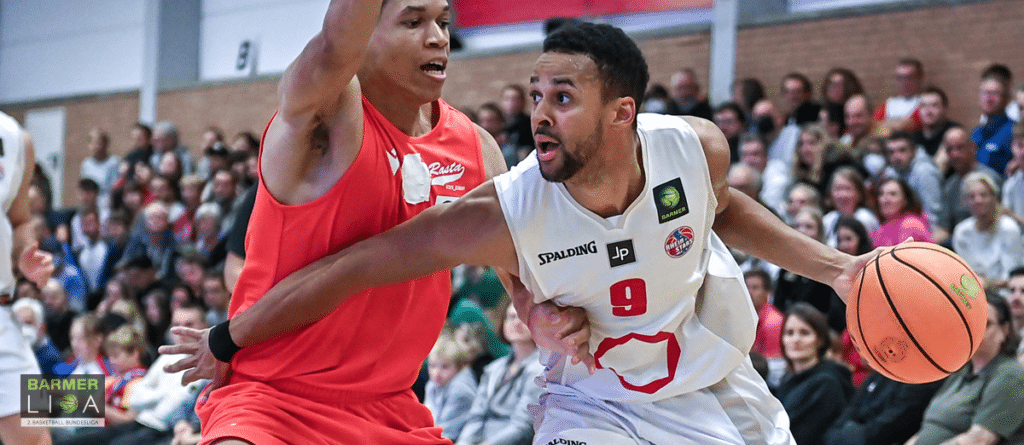 Basketballspiel der RheinStars Köln: Spieler Nummer 9 im weißen Trikot dribbelt gegen Gegenspieler in Rot.