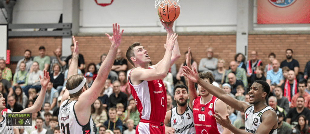 Basketballspieler der RheinStars Köln wirft Ball zum Korb, Zuschauer im Hintergrund.