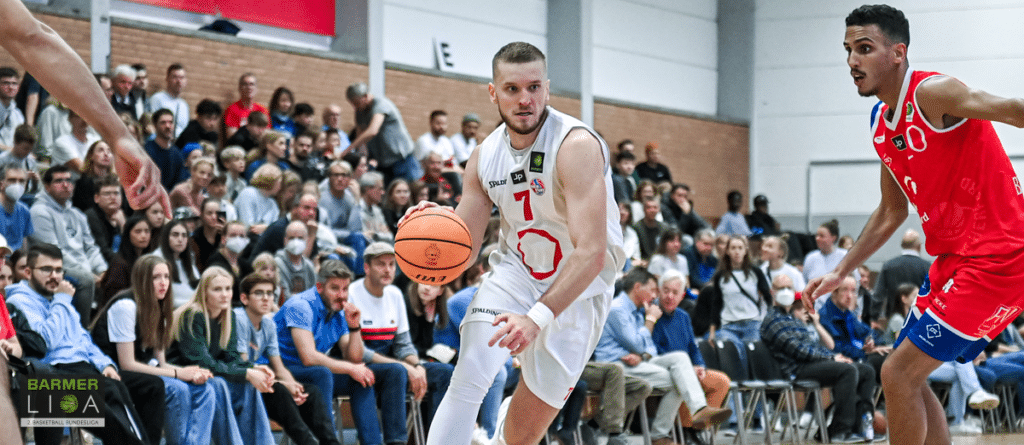 Basketballspieler der RheinStars Köln im weißen Trikot dribbelt gegen roten Gegner.