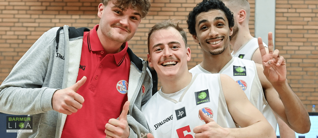 Vier Basketballspieler der RheinStars Köln posieren lachend vor einer Backsteinwand.