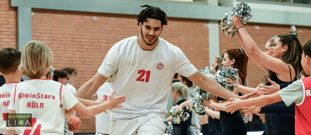 Basketballspieler der RheinStars Köln wird von Cheerleadern mit Pompons begrüßt.