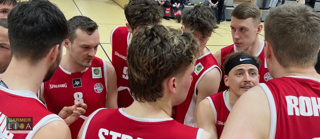 Basketballmannschaft RheinStars Köln in roten Trikots beim Mannschaftshuddel in der Sporthalle.