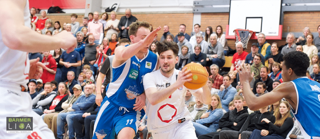 Basketballspiel der RheinStars Köln: Spieler in weißem Trikot dribbelt gegen Gegner in blauem Trikot vor gefüllter Zuschauerhalle.