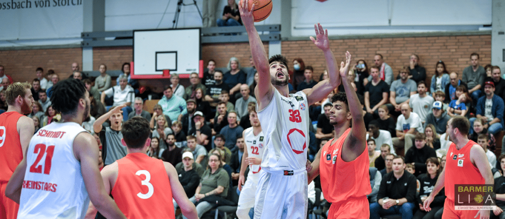 Basketballspiel der RheinStars Köln: Spieler in Weiß wirft Ball, Gegner in Rot verteidigt, Zuschauer im Hintergrund.