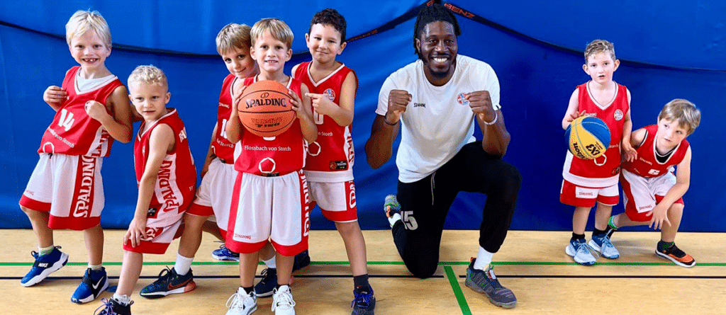 Ein erwachsener Basketballtrainer posiert mit sieben Kindern in RheinStars-Köln-Trikots in einer Sporthalle.