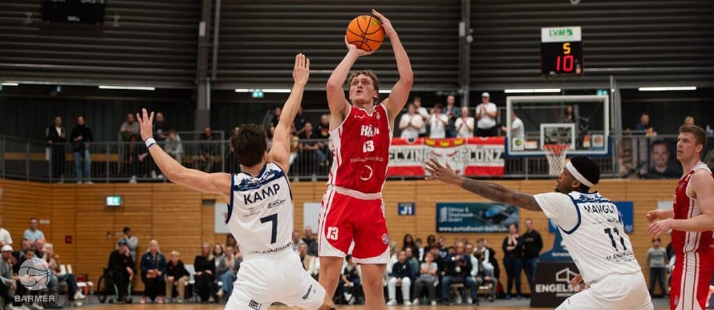 Basketballspiel der RheinStars Köln: Spieler im roten Trikot wirft Ball über zwei verteidigende Gegner im weißen Trikot.