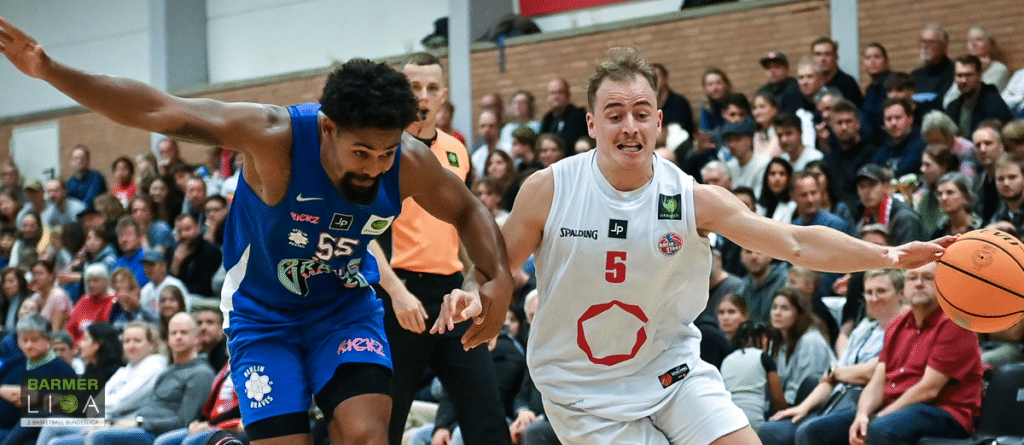 Basketball-Spieler der RheinStars Köln in weißem Trikot mit Nummer 5 dribbelt gegen blauen Gegenspieler.