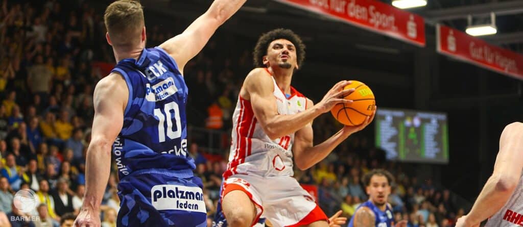 Basketballspieler der RheinStars Köln in rotem Trikot dribbelt gegen Gegner in blauem Trikot.