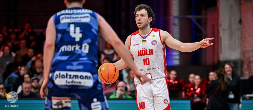 Basketballspieler der RheinStars Köln in weißem Trikot mit Nummer 10 dribbelt gegen blauen Gegner.