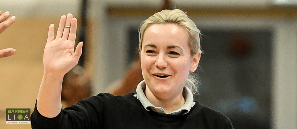 Frau mit blondem Haar winkt lächelnd in einer Sporthalle, BARMER LIGA Logo sichtbar.