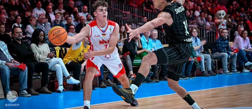 RheinStars-Spieler in rotem Trikot dribbelt Basketball gegen schwarzgekleideten Gegenspieler vor vollem Publikum.