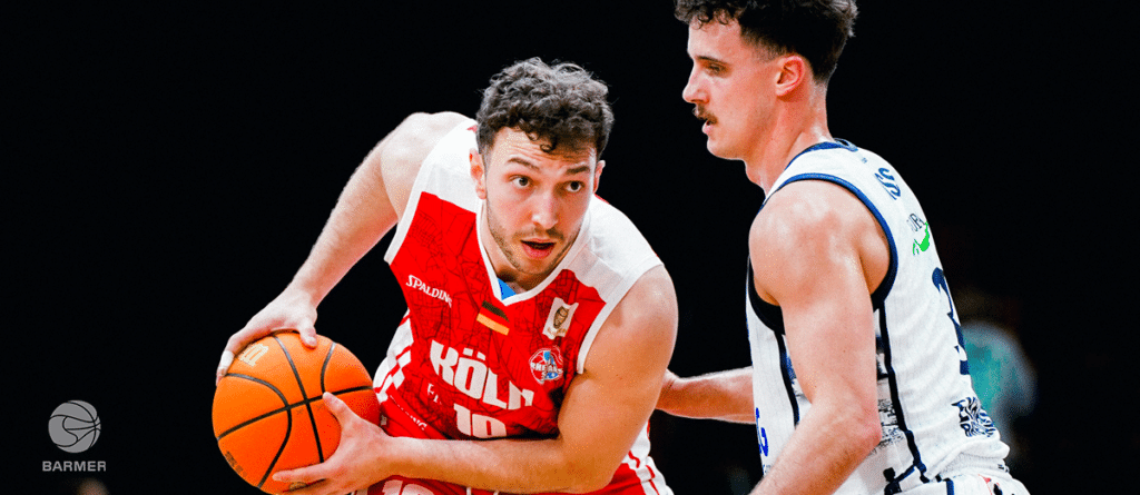Basketballspieler der RheinStars Köln in rotem Trikot dribbelt gegen Gegner in weißem Trikot.