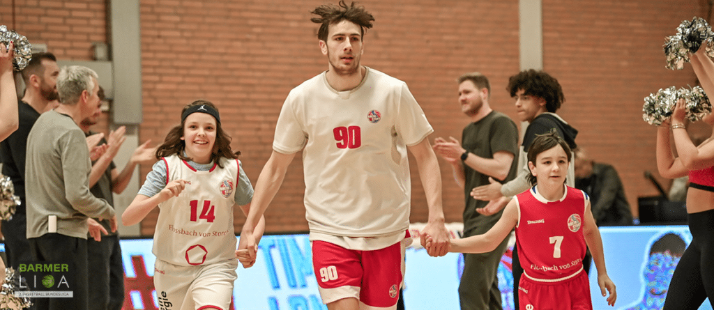 Ein erwachsener Spieler der RheinStars Köln läuft Hand in Hand mit zwei jungen Kindern in Trikots über das Spielfeld, umgeben von applaudierenden Zuschauern.