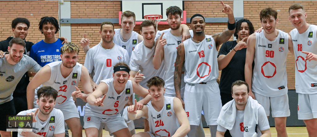 RheinStars Köln Basketballmannschaft posiert lachend in weißen Trikots in einer Sporthalle.