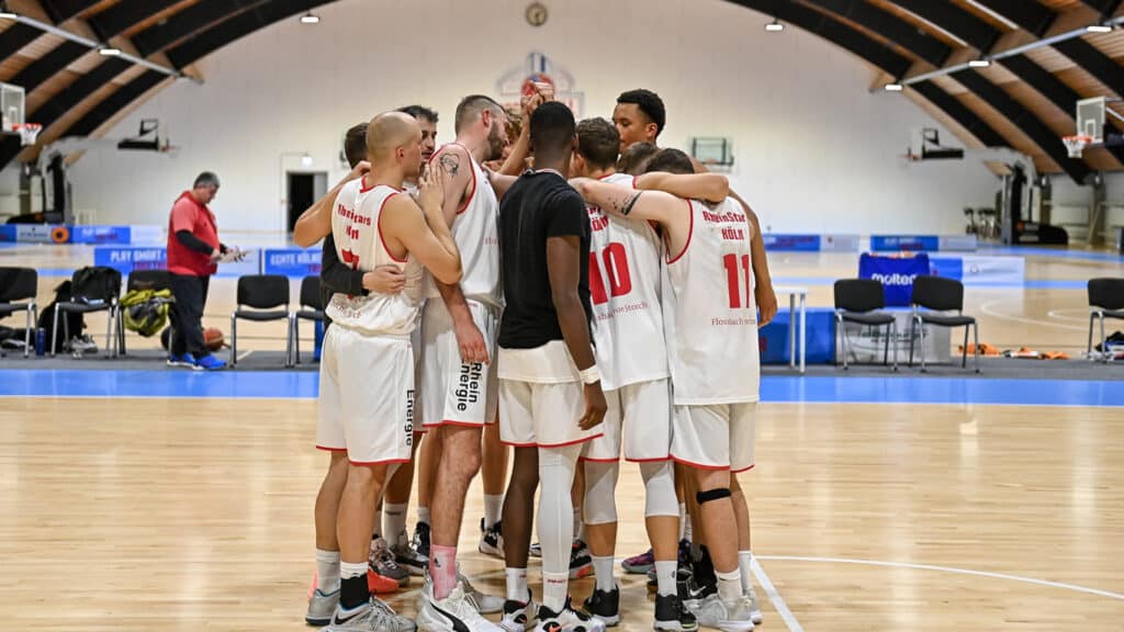 Basketballmannschaft der RheinStars Köln im Teamkreis auf dem Spielfeld einer Sporthalle.