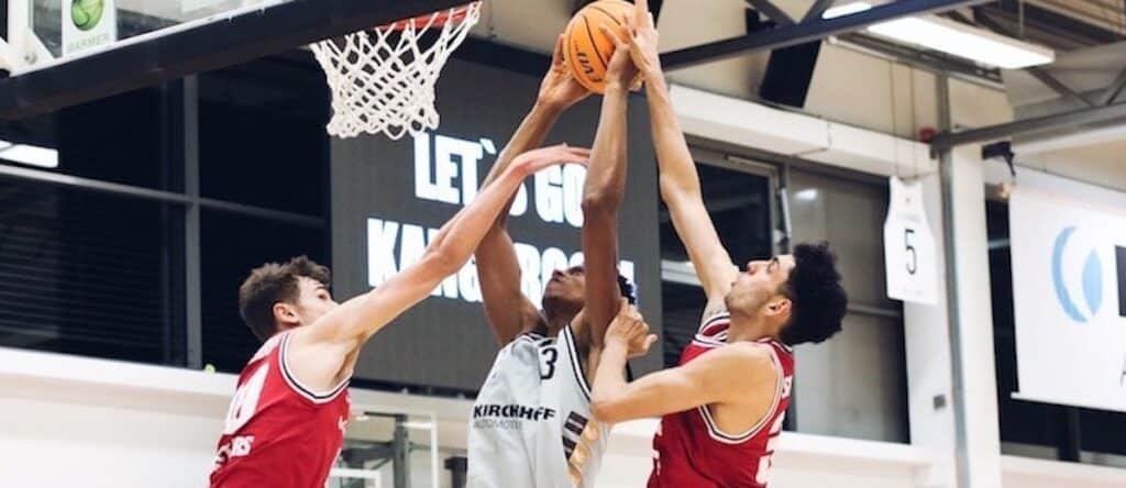 Basketballspiel der RheinStars Köln: Spieler in Weiß springt zwischen zwei Gegnern in Rot nach dem Ball.