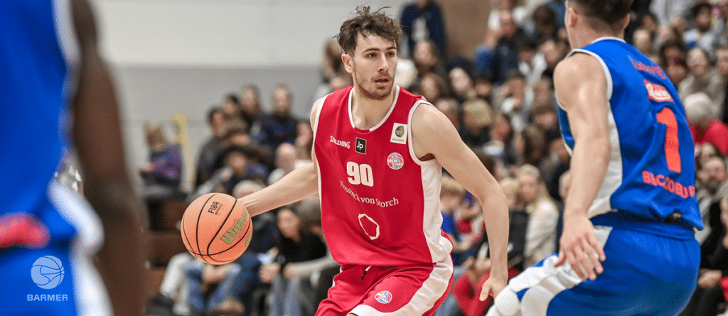 Basketballspieler der RheinStars Köln in rotem Trikot dribbelt gegen blaue Gegenspieler.
