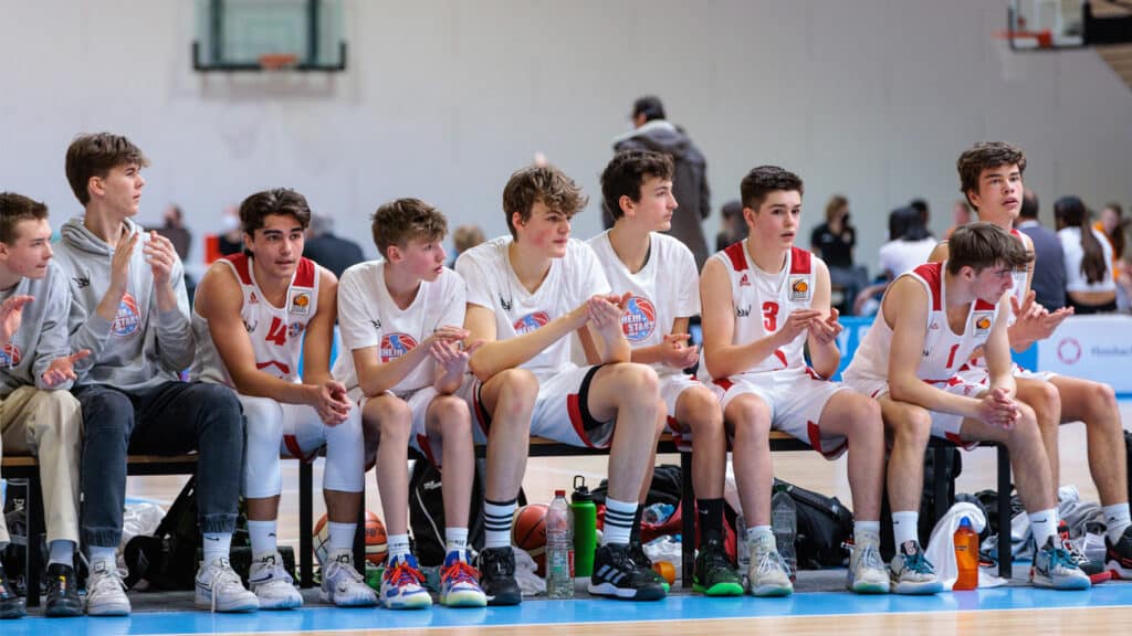 Jugendbasketballmannschaft der RheinStars Köln sitzt auf der Ersatzbank einer Sporthalle.