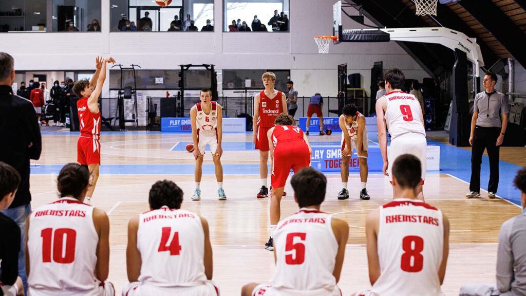 Basketballspiel der RheinStars Köln: Spieler in roten und weißen Trikots auf dem Spielfeld einer Indoor-Halle.