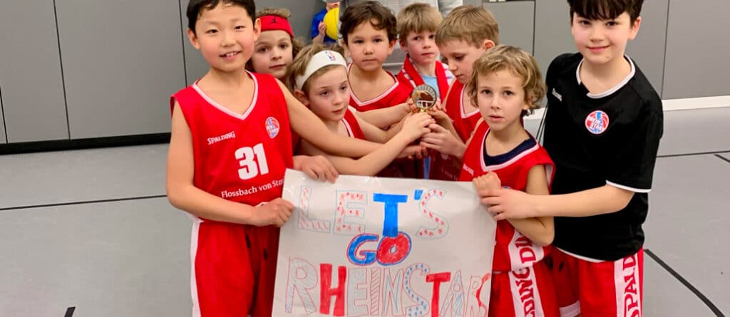 Gruppe von Kindern der RheinStars Köln hält stolz ihr selbstgemaltes Teamplakat in einer Sporthalle.