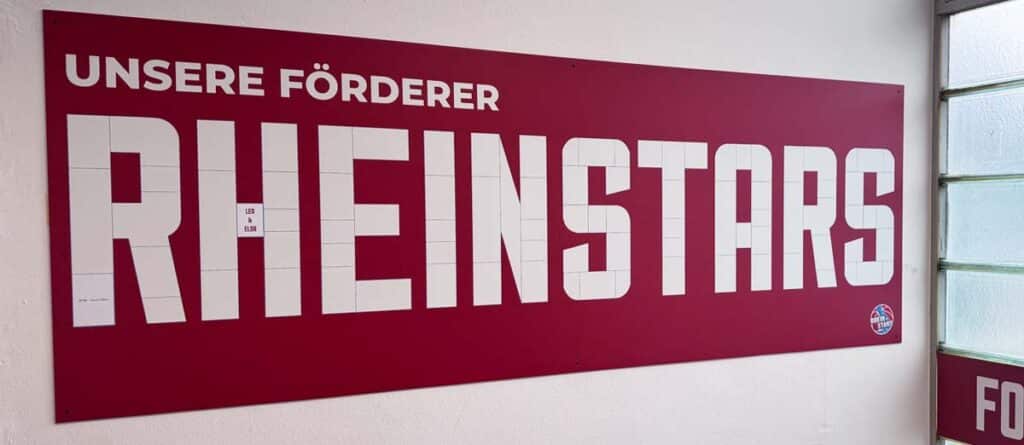 Großes rotes Wandschild mit weißer Aufschrift "UNSERE FÖRDERER" und "RHEINSTARS" in einer Sporthalle der RheinStars Köln.