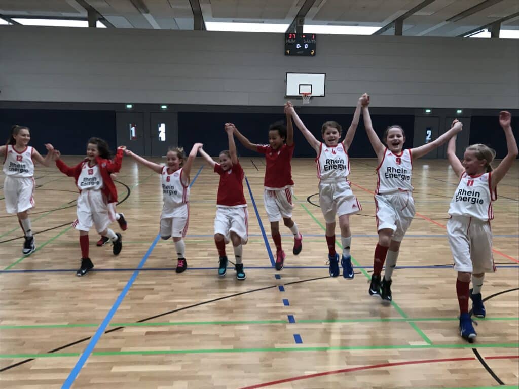 Junge Basketballspielerinnen der RheinStars Köln springen jubelnd in einer Sporthalle.
