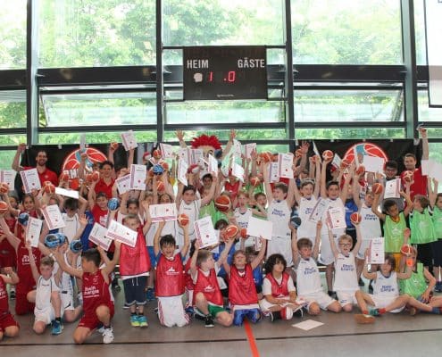 Große Gruppe von Kindern der RheinStars Köln in rot-weißen Trikots posiert vor einer Sporthallenwand mit Anzeigetafel.